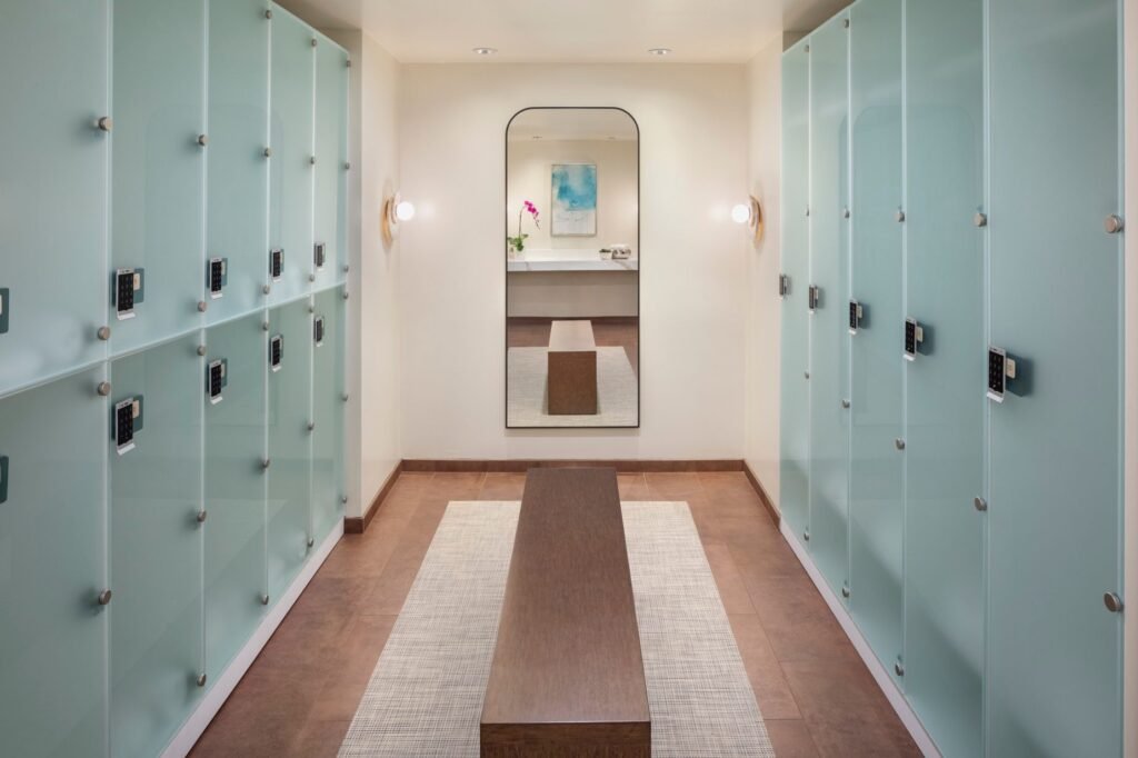 A modern spa locker room corridor lined on both sides with frosted glass lockers featuring digital keypads. A long wooden bench sits on a light woven rug over dark tiled floors in the center of the walkway. At the far end, a large arched mirror reflects a small vanity with an orchid and a piece of abstract blue wall art.