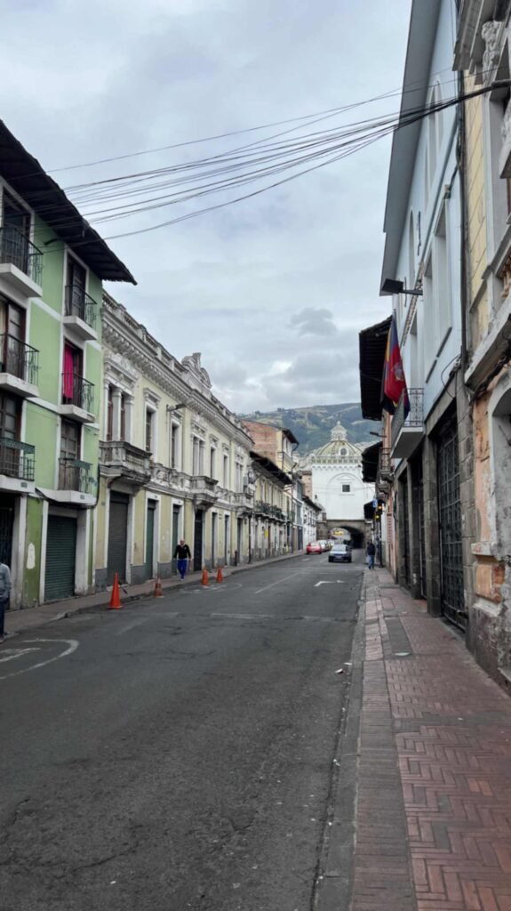 Street level view looking down a narrow colonial road in Quito lined with multi story historic buildings featuring iron balconies and the stone Arch of Santo Domingo framing a church dome in the far distance