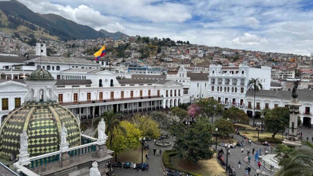 High angle view of Plaza de la Independencia in Quito Ecuador featuring historic white colonial buildings with arched walkways and a prominent green and yellow tiled dome in the foreground under a cloudy sky with hills rising in the background