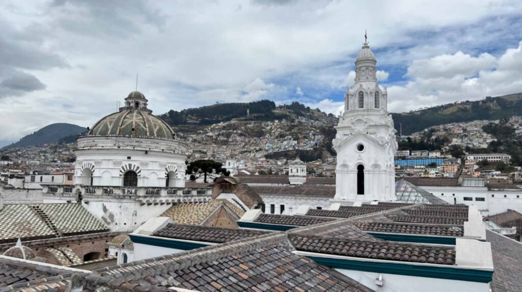 Rooftop view of the Quito Metropolitan Cathedral showcasing a large green and brown tiled dome and an ornate white bell tower against a hillside city backdrop under a cloudy sky