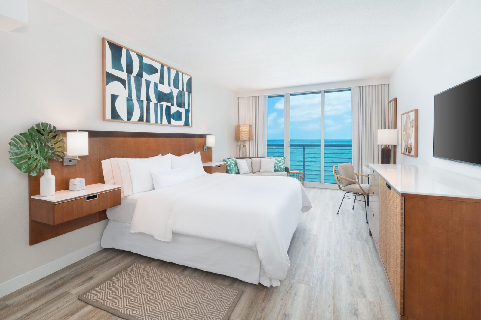 A bright hotel guest room with a view of the blue ocean through large sliding glass doors. The room features a king bed with crisp white linens, a wooden headboard, floating nightstands, and a large abstract blue and white art piece on the wall. A wooden dresser with a flat screen television faces the bed, and a small seating area sits near the balcony.
