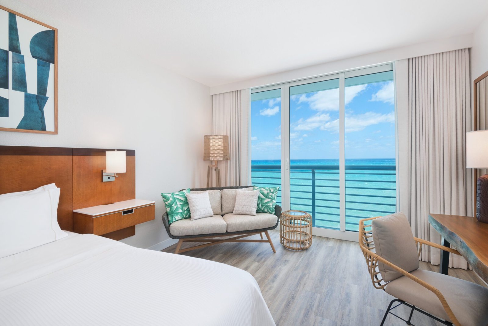 A bright hotel room corner featuring a small woven sofa with teal and white pillows next to floor-to-ceiling glass sliding doors. Outside the balcony railing, a wide expanse of turquoise and blue ocean meets a partly cloudy sky. The room includes light wood flooring, a wooden desk with a woven chair on the right, and the corner of a bed with white linens on the left.