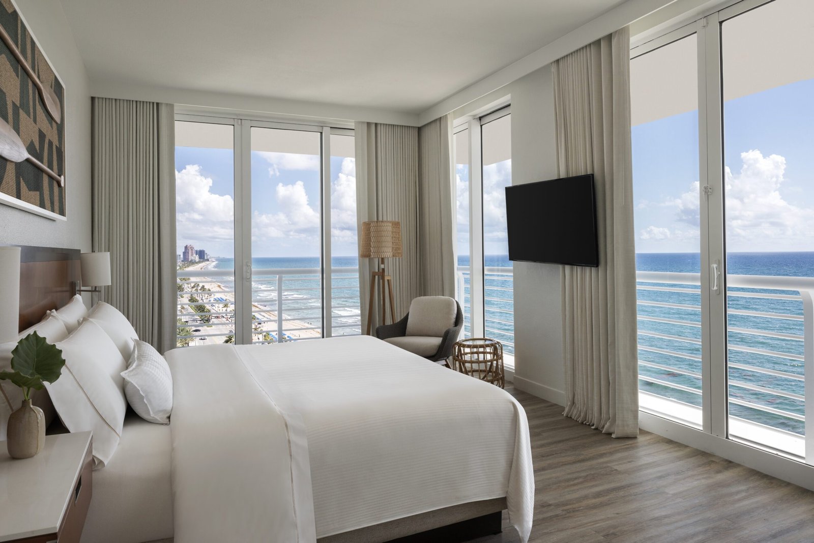 A spacious hotel guest room with multiple floor-to-ceiling glass doors showcasing wrap-around views of the sandy beach and ocean. The room features a king bed with crisp white bedding, an armchair nestled in the corner by a woven floor lamp, and a flat screen television mounted on the wall. Light grey curtains frame the bright windows under a partly cloudy sky.