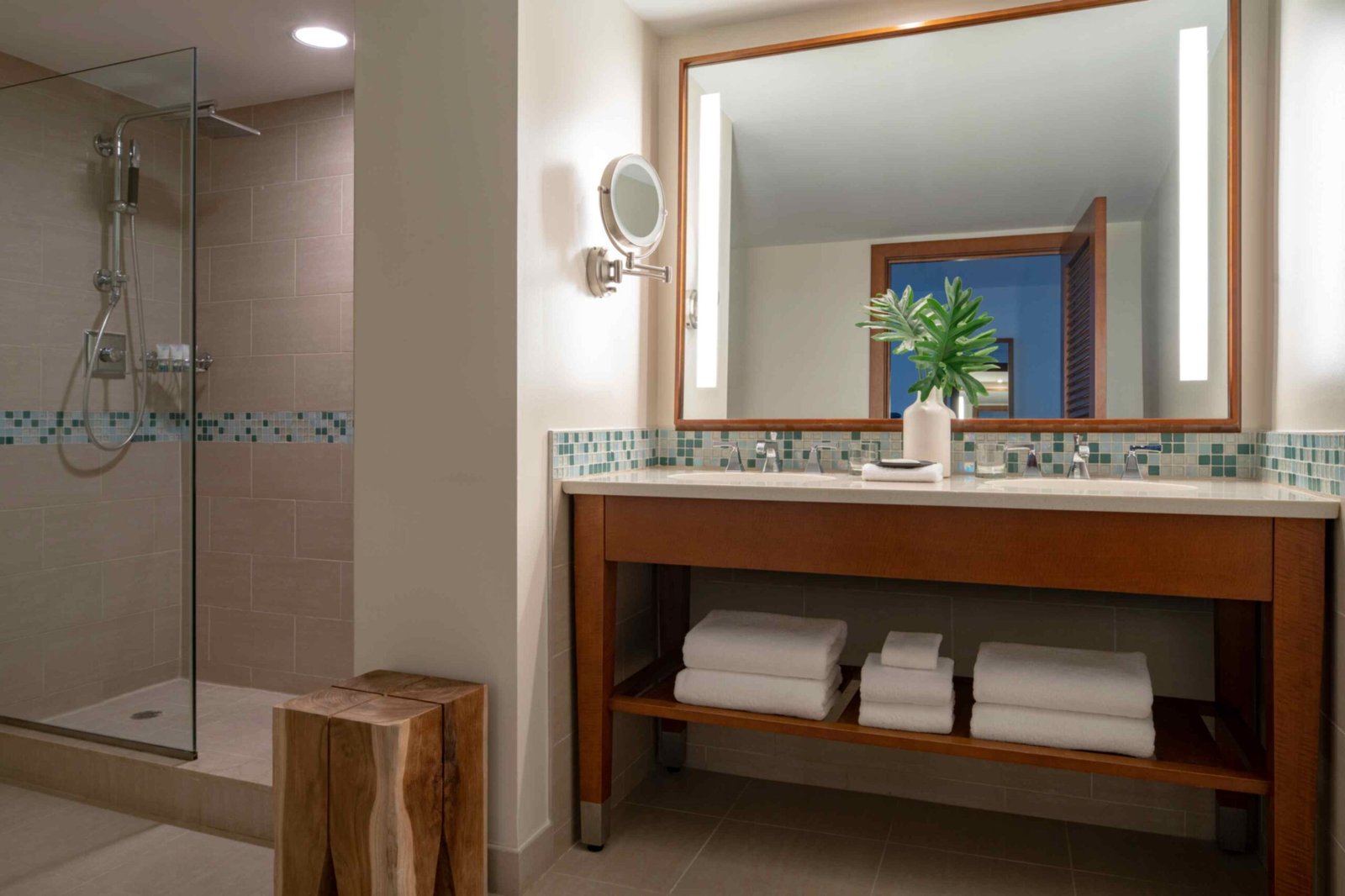 A modern hotel bathroom featuring a wooden double vanity with a light countertop and a large illuminated mirror. To the left, a walk in shower enclosed in glass displays beige tiles with a blue and green mosaic accent strip. Rolled white towels rest on the lower shelf of the vanity, and a rustic wooden block stool sits on the tiled floor.