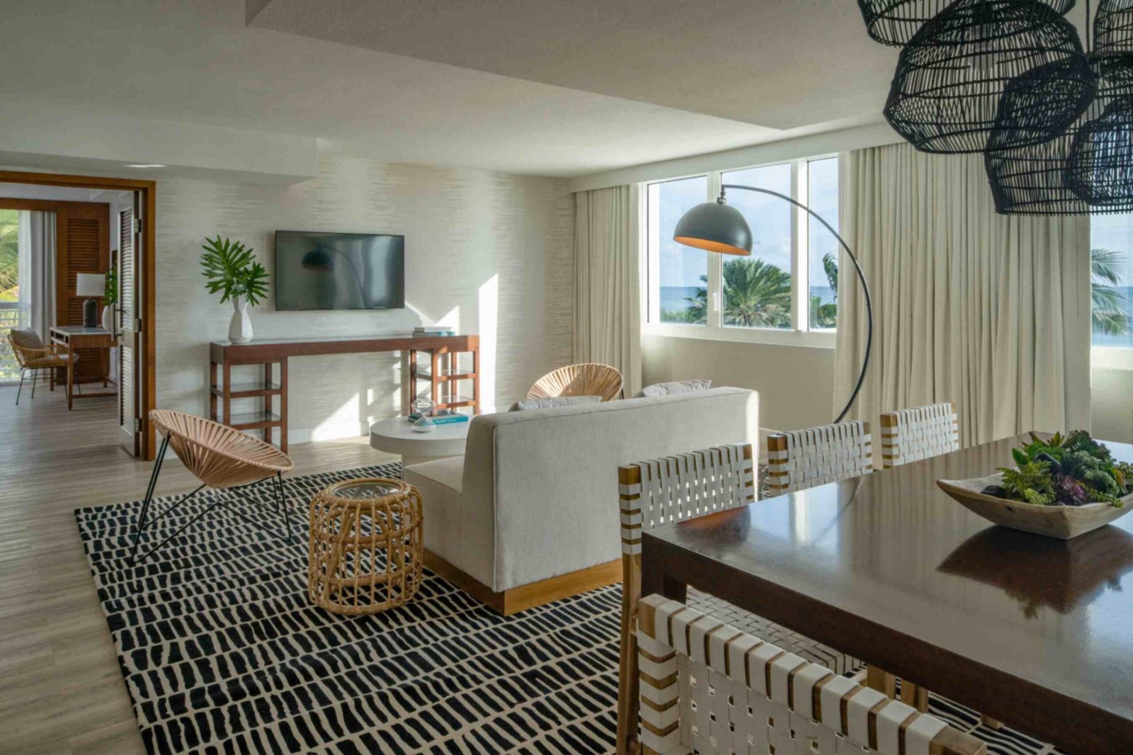 A spacious hotel suite living and dining area with a modern coastal design. The room features a beige sofa on a bold black and white patterned rug, woven accent chairs, a wooden console table under a wall mounted television, and a large arched floor lamp. On the right, a wooden dining table is surrounded by woven chairs beneath dark wicker pendant lights, with bright windows showing palm trees outside.