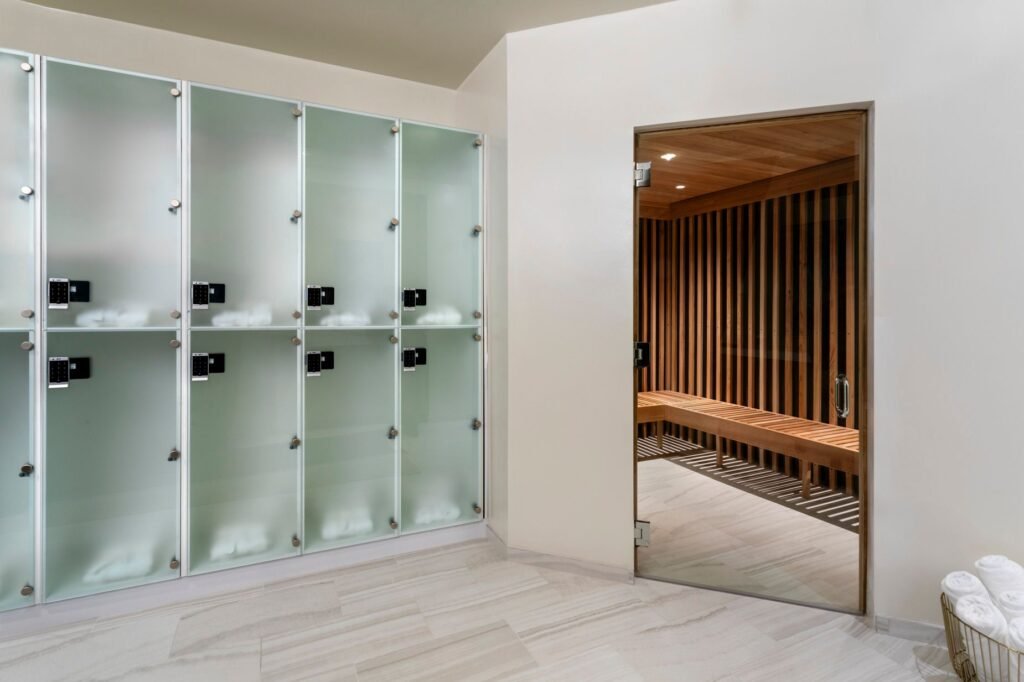 A bright spa locker room area with light marble tile flooring and frosted glass lockers featuring black digital locks. On the right, a glass door opens into a wooden sauna equipped with slatted timber walls and a long matching wood bench. A basket filled with rolled white towels rests in the lower right corner.