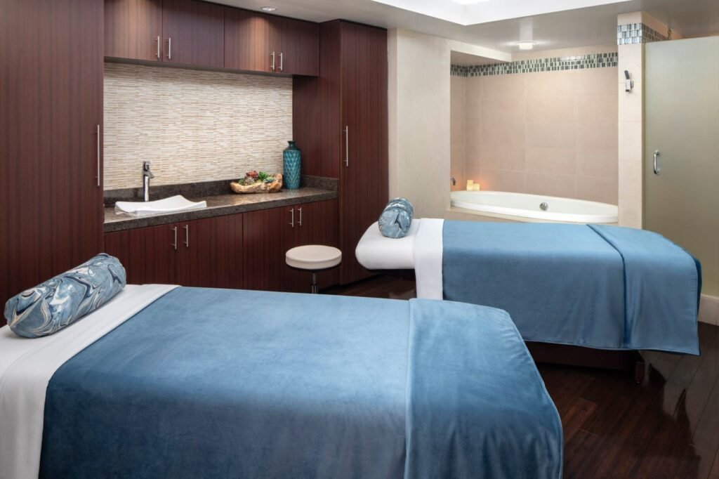 A peaceful couples spa treatment room with dark wood flooring and cabinetry. Two massage tables dressed in plush blue blankets and patterned bolster pillows stand in the foreground. In the background, a small vanity counter features a modern vessel sink, while a separate alcove holds a large white soaking bathtub illuminated by lit candles.