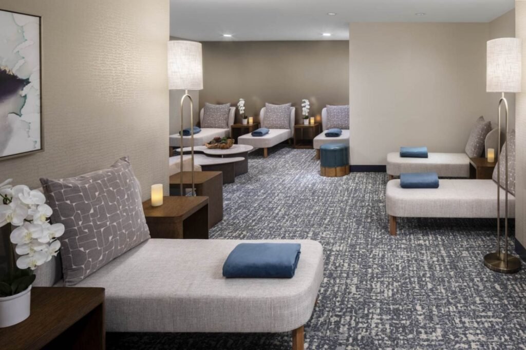 A spacious hotel spa relaxation room with a textured grey and white carpet and soft lighting. Several cushioned chaise lounges with patterned pillows and folded blue blankets line the walls. Wooden side tables with lit candles and white orchids sit next to the lounges alongside tall metal floor lamps with fabric shades.
