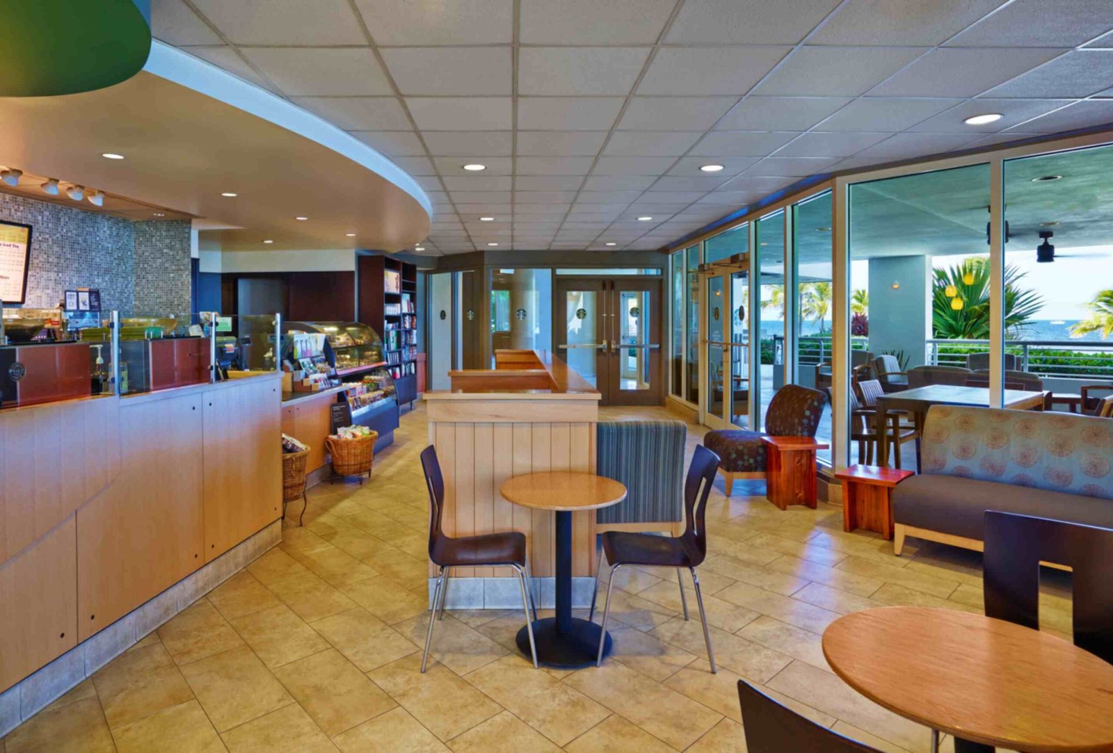 An interior view of a bright resort cafe and marketplace featuring a curved wooden service counter and a small dining area with tables and chairs. Large floor-to-ceiling windows look out onto an outdoor covered patio with ocean views and palm trees.