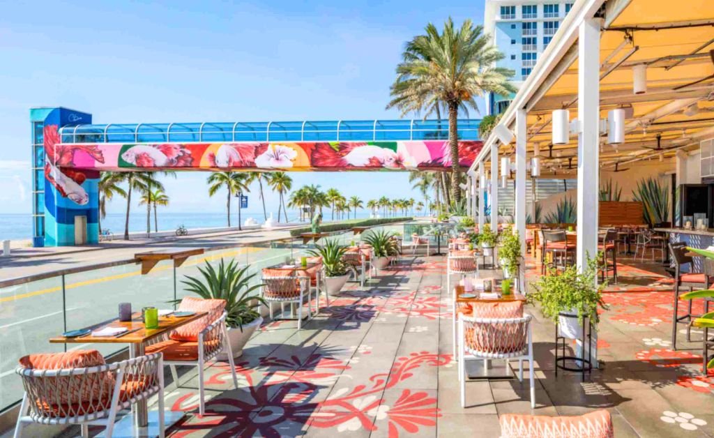 An open air restaurant patio overlooking a coastal road and the ocean. The patio features patterned floor tiles, modern dining tables, woven chairs with orange cushions, and potted plants. In the background, an elevated glass skywalk bridge decorated with vibrant red and pink floral art stretches toward the beach. Palm trees line the sandy shoreline under a bright blue sky.