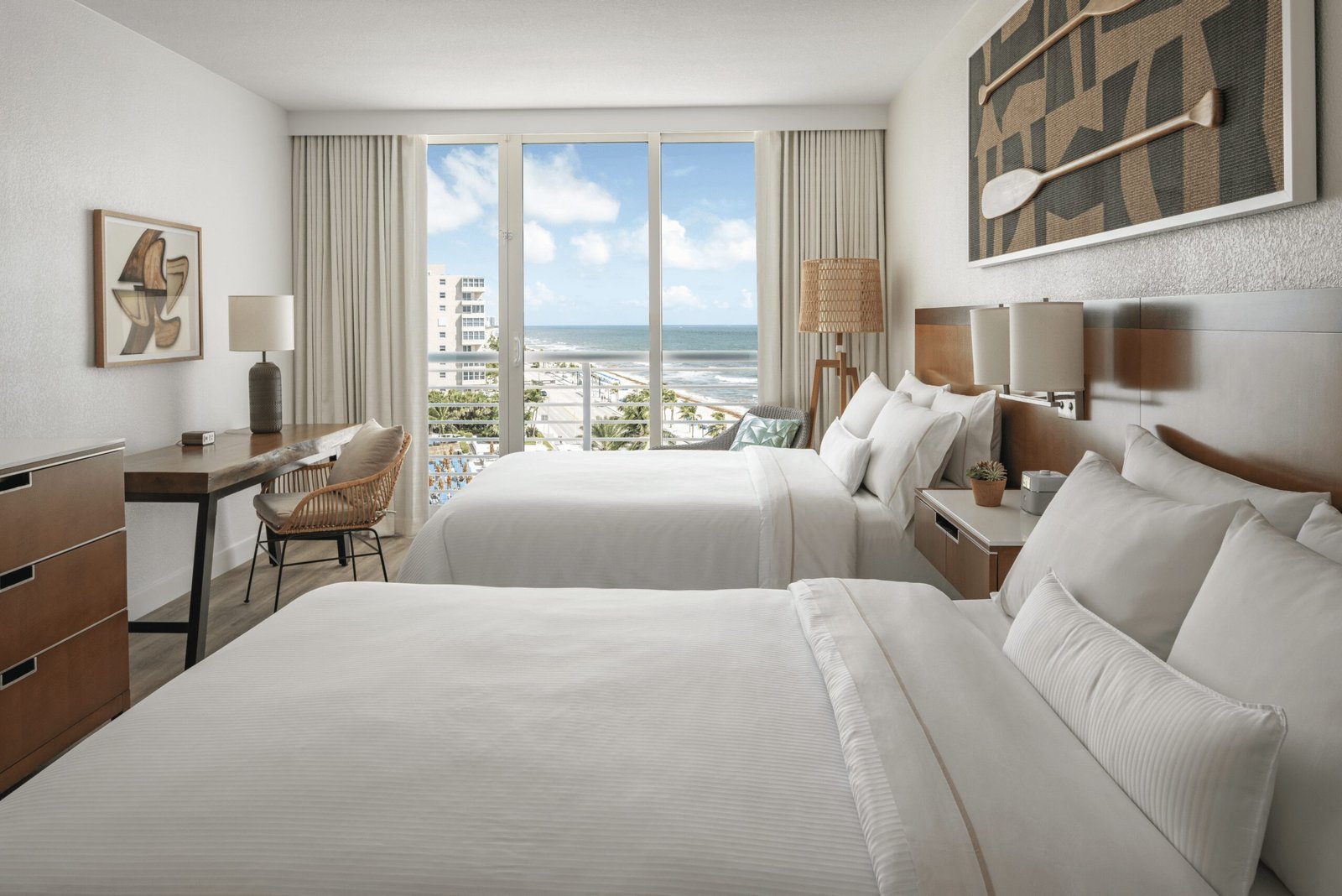 A bright hotel guest room featuring two queen beds with crisp white bedding. On the right, a wooden headboard is topped by framed art displaying decorative wooden oars. On the left, a wooden writing desk with a woven chair stands against the wall. Large glass sliding doors in the back open to a balcony with views of the beach and ocean under a blue sky.