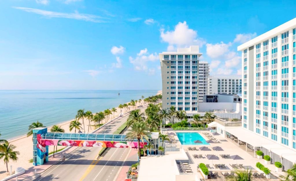 An elevated high angle view of a coastal resort pool deck and the adjacent beach. On the right, the multi story hotel building overlooks an outdoor swimming pool surrounded by gray lounge chairs and umbrellas. A coastal road runs between the resort and the sandy beach, spanned by an elevated blue skywalk bridge with vibrant floral art. Palm trees line the road and shoreline under a clear blue sky.