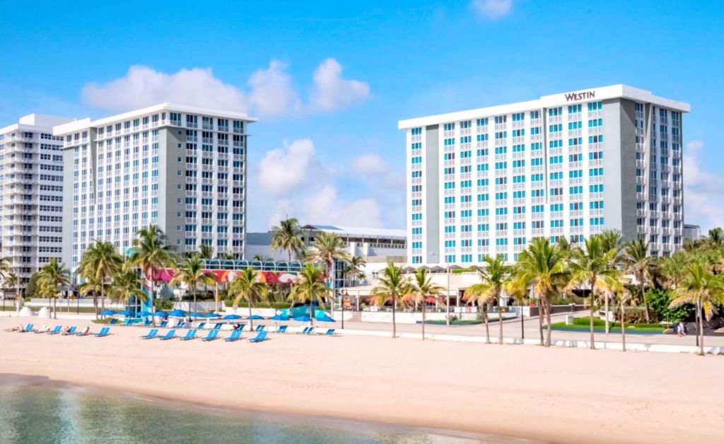 A wide view of The Westin Fort Lauderdale Beach Resort towers standing along a sandy beach. The modern white multi story hotel buildings face a shoreline lined with palm trees, blue beach umbrellas, and matching blue lounge chairs. Calm, clear ocean water ripples in the foreground beneath a bright blue sky with light clouds.