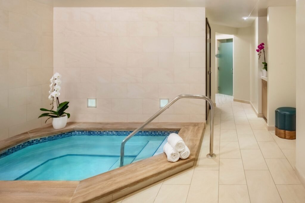 An indoor resort spa hot tub area with beige tiled walls and flooring. The sunken whirlpool is framed by a wood-grain border and blue mosaic tiles, featuring clear turquoise water and a metal handrail. Three rolled white towels sit on the edge near a potted white orchid, while a hallway leads away in the background.